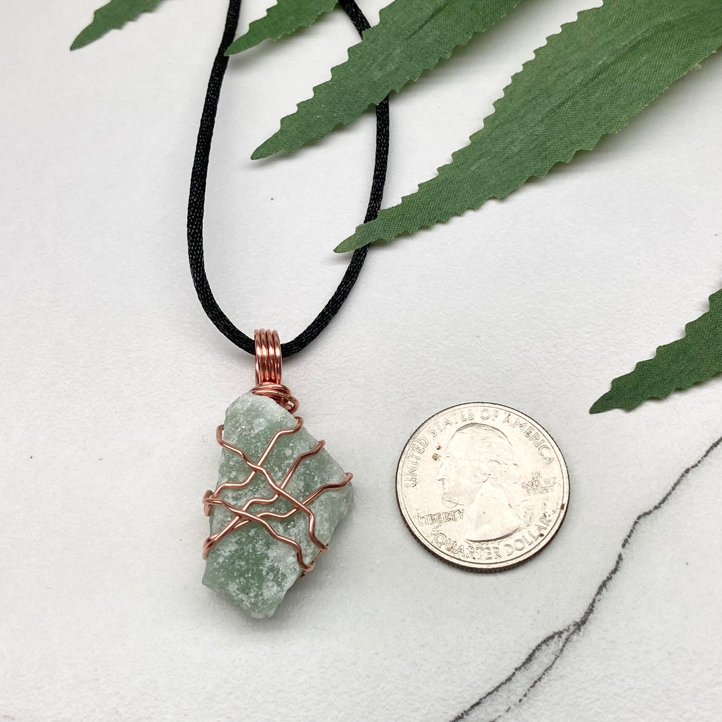 Necklace with a green stone pendant next to a coin on a marble surface with leaves in the background