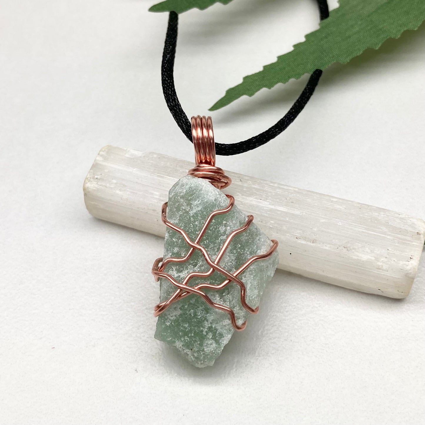 Green crystal pendant with copper wire wrapping on a black cord, placed on a marble surface with green leaves.