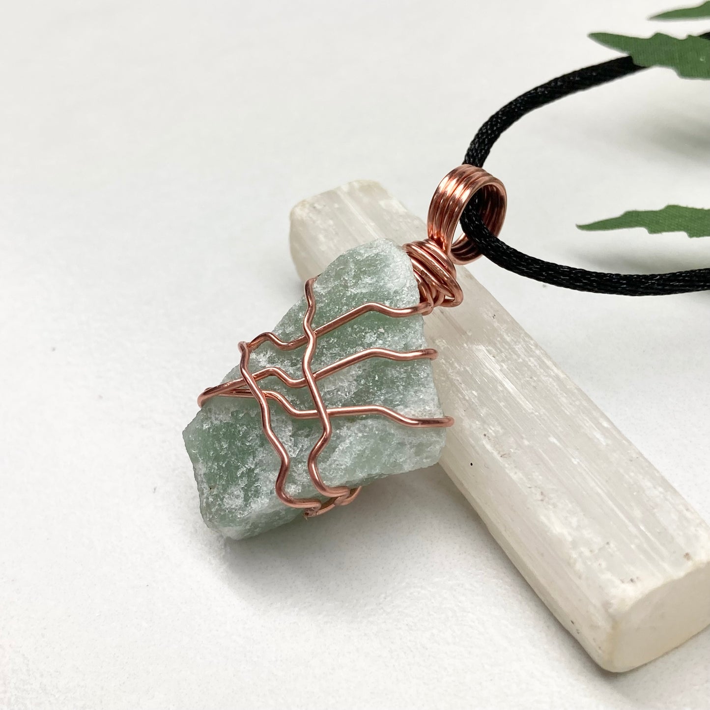 Necklace with a green stone pendant and copper wire wrapping, placed on a white surface with a crystal and leaf in the background.