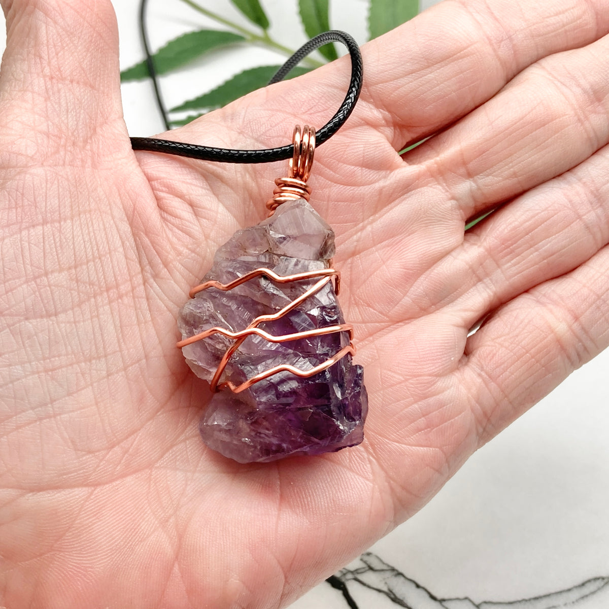 A raw purple amethyst crystal pendant wrapped with copper wire, hanging from a black cord and held in a person's hand.