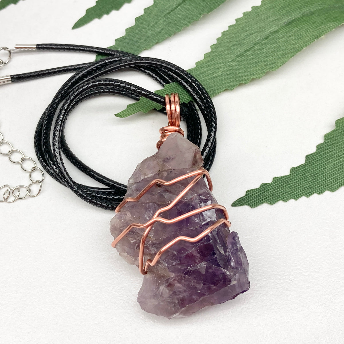 A raw amethyst crystal pendant wrapped in copper wire, hanging from a black cord necklace with a silver clasp, displayed on a white background with green leaves.