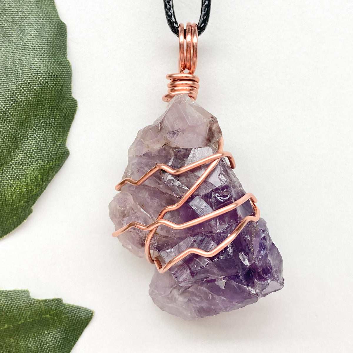 A large raw amethyst crystal wrapped in copper wire, hanging from a black cord, displayed on a white background with a green leaf on the left.