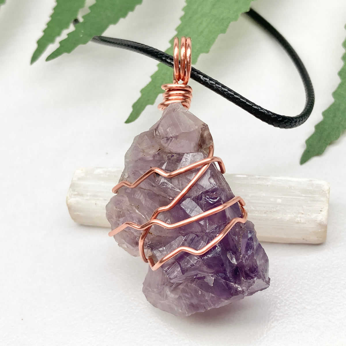 A raw amethyst crystal pendant wrapped with copper wire, hanging from a black cord, displayed on a white selenite rod with green leaves in the background.