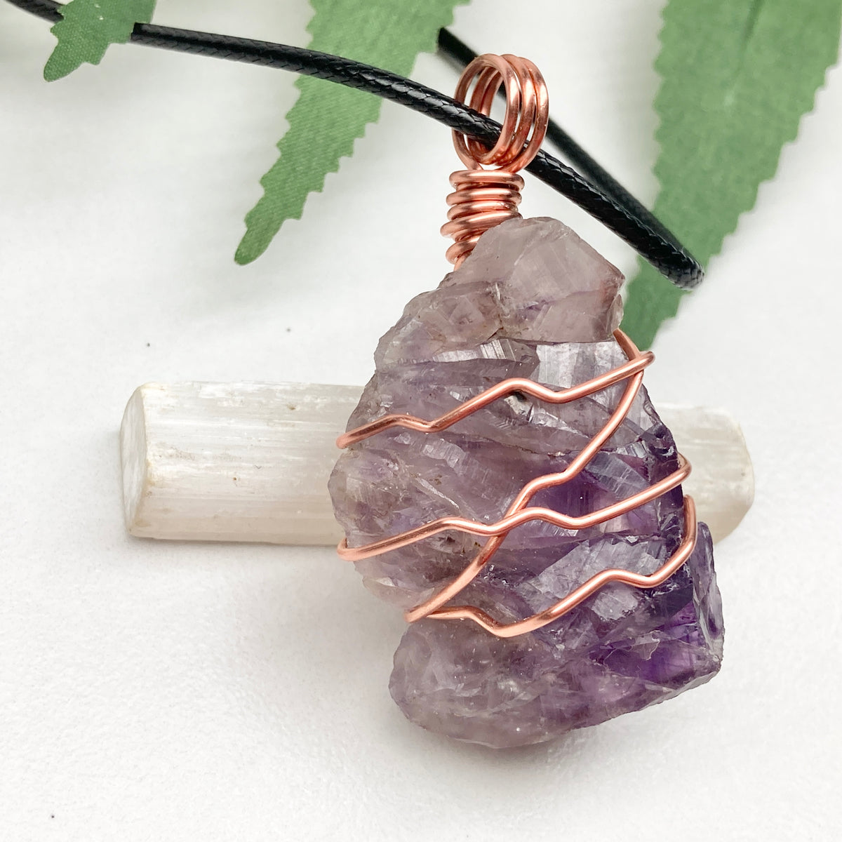 A raw amethyst crystal pendant wrapped in copper wire, hanging from a black cord, displayed on a white selenite wand against a white background with green leaves.