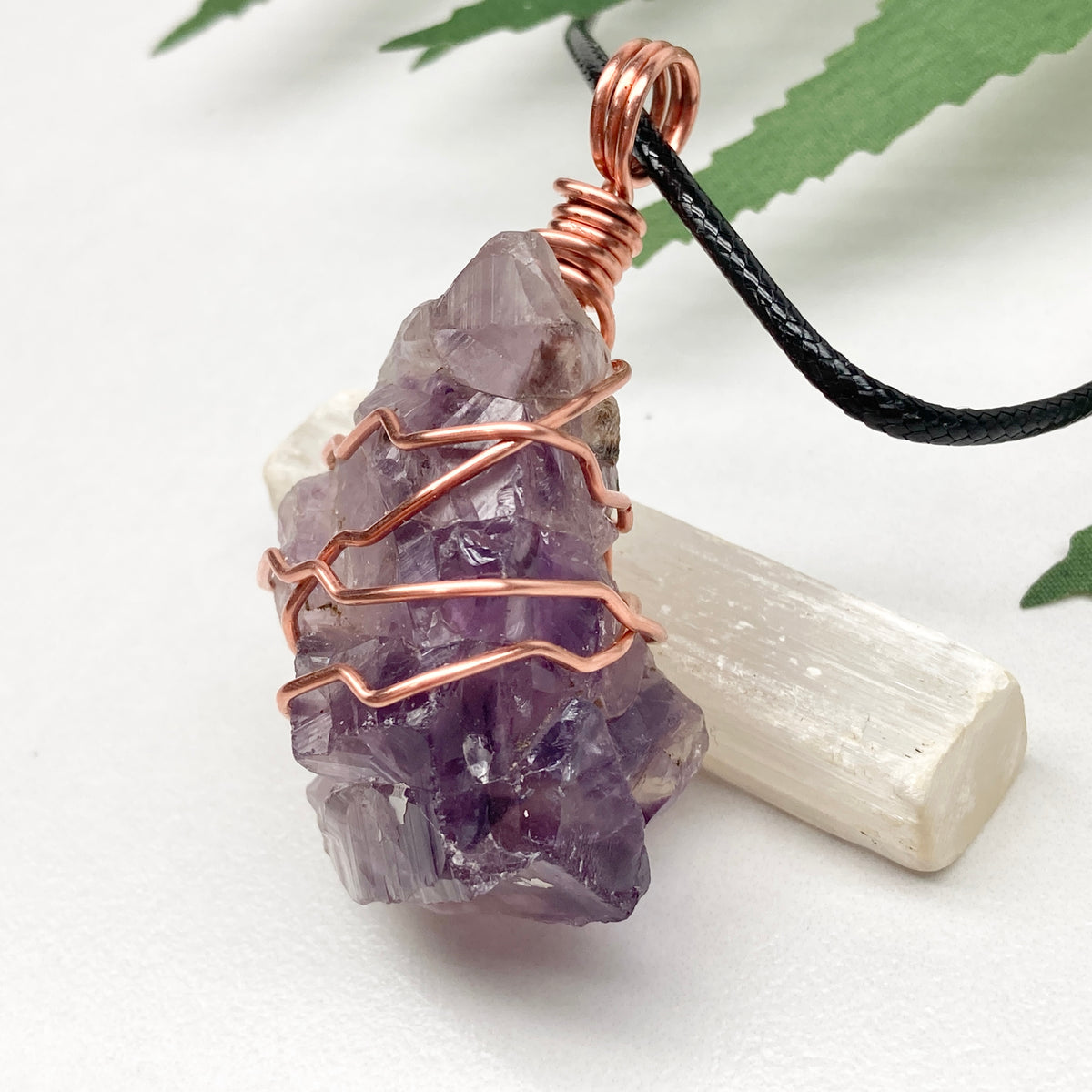 A raw purple amethyst crystal cluster pendant wrapped in copper wire, hanging from a black cord, displayed on a white crystal stick against a white background with green leaves.