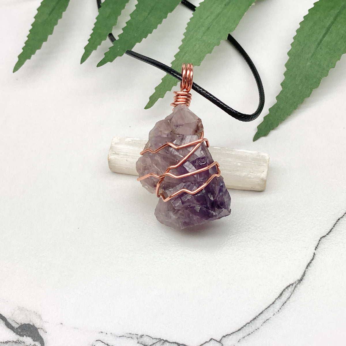 A raw amethyst crystal pendant necklace wrapped in copper wire, resting on a white selenite wand against a white marble background with green leaves.
