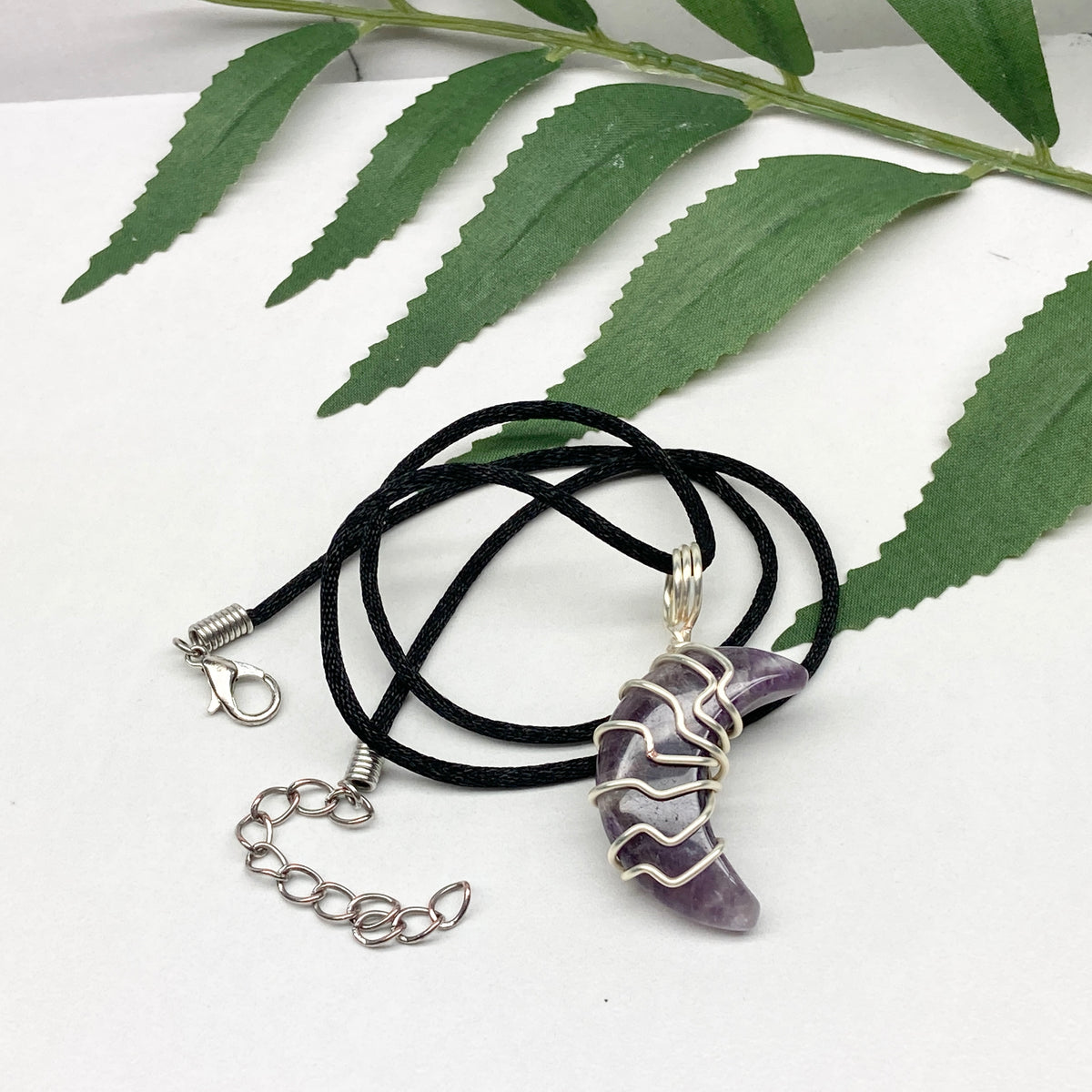 Purple Amethyst crescent moon necklace wrapped in silver-colored copper wire and hanging on a black chain. Handmade during the Moon in Leo.