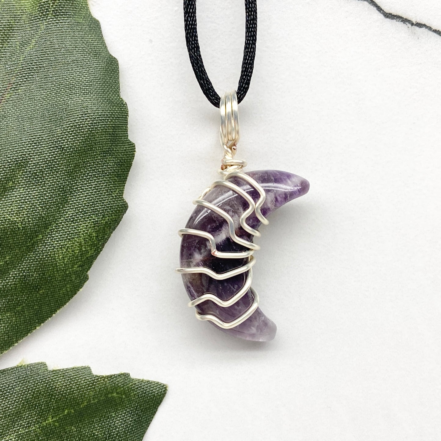 Purple Amethyst crescent moon necklace wrapped in silver-colored copper wire and hanging on a black chain. Handmade during the Moon in Leo.