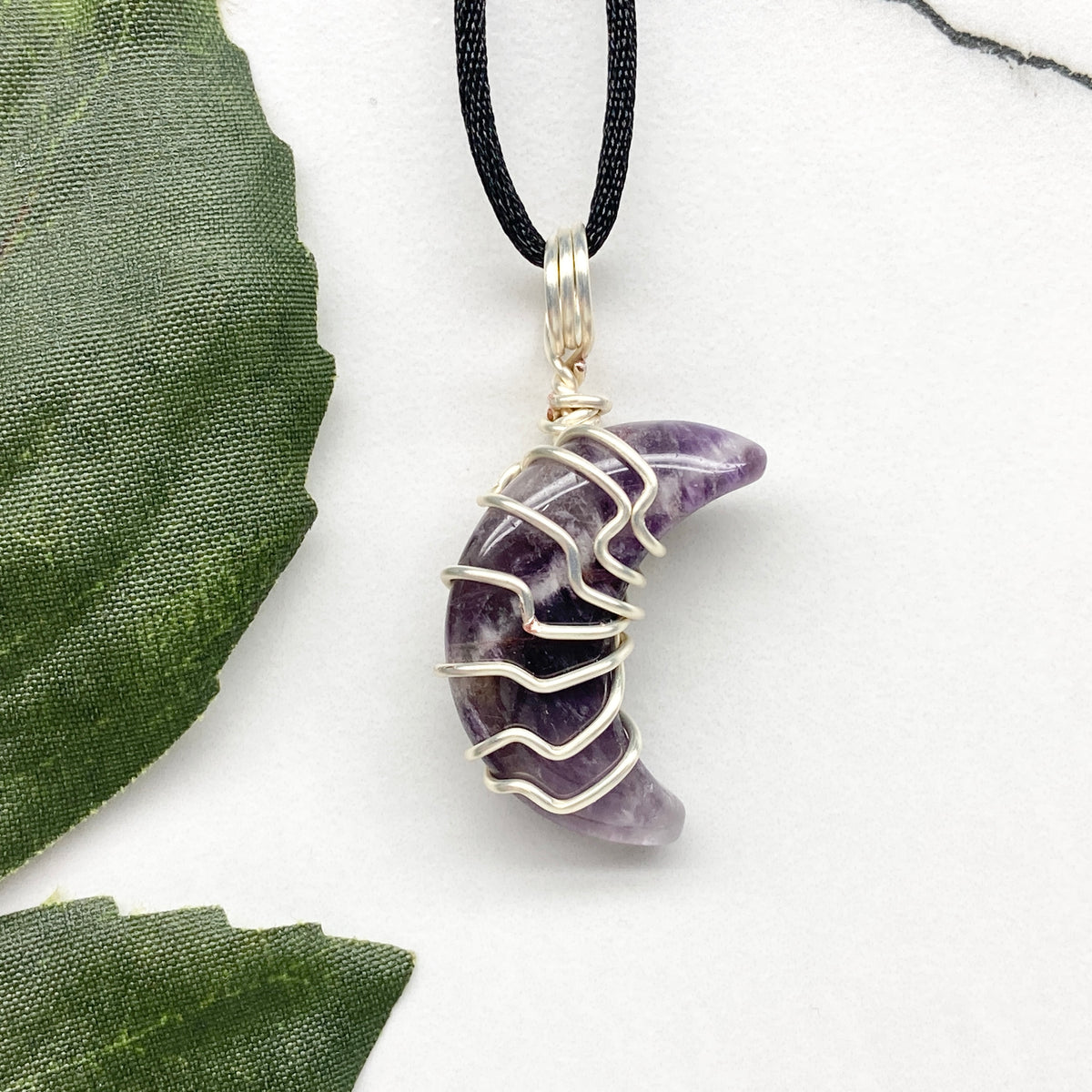 Purple Amethyst crescent moon necklace wrapped in silver-colored copper wire and hanging on a black chain. Handmade during the Moon in Leo.