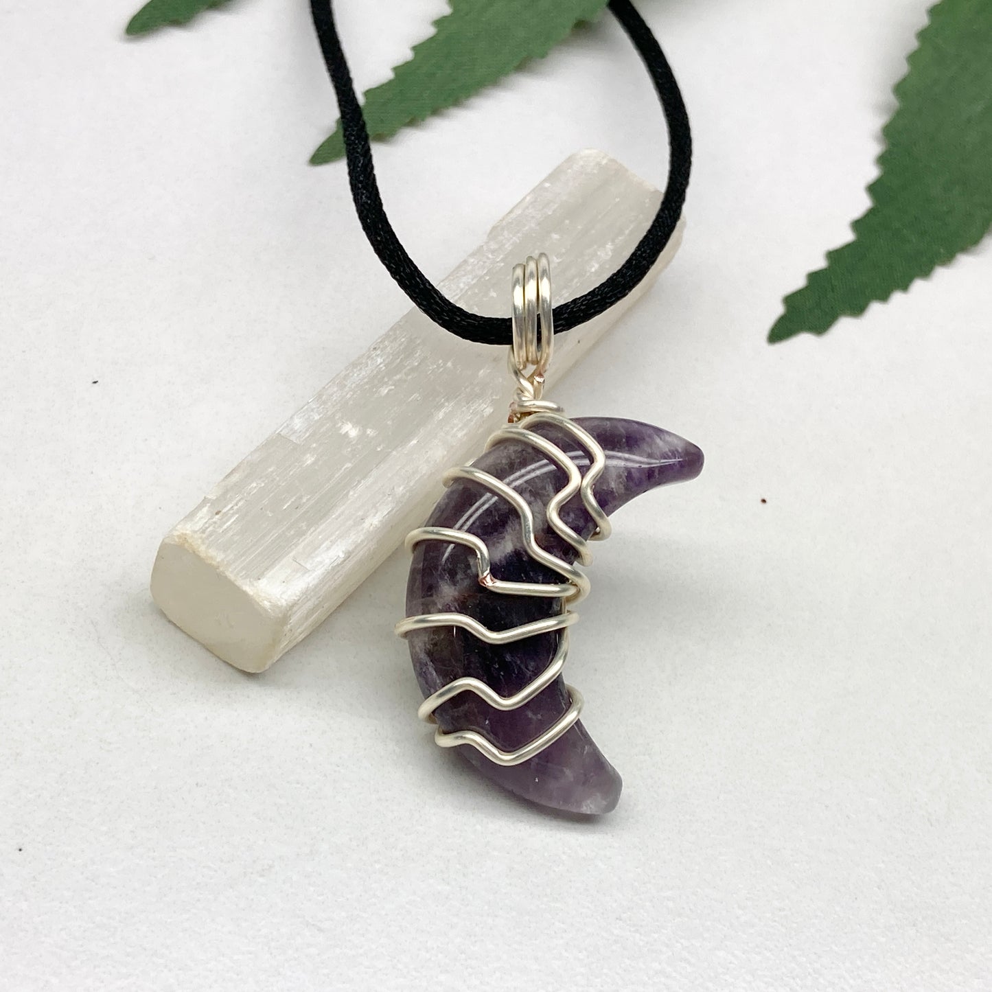 Purple Amethyst crescent moon necklace wrapped in silver-colored copper wire and hanging on a black chain. Handmade during the Moon in Leo.