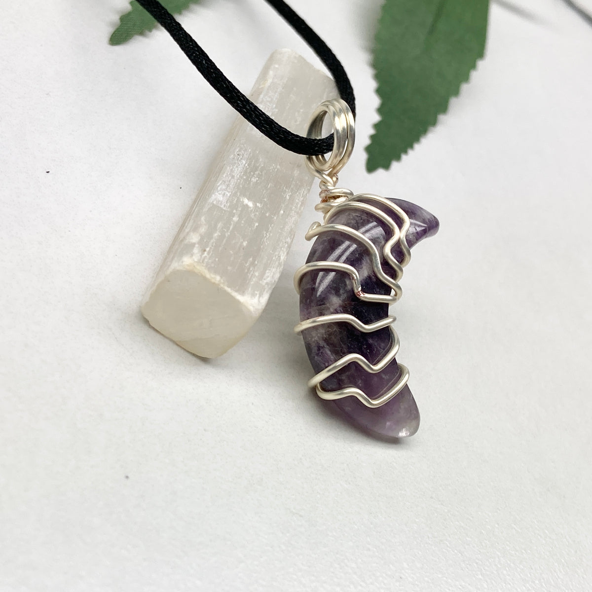 Purple Amethyst crescent moon necklace wrapped in silver-colored copper wire and hanging on a black chain. Handmade during the Moon in Leo.