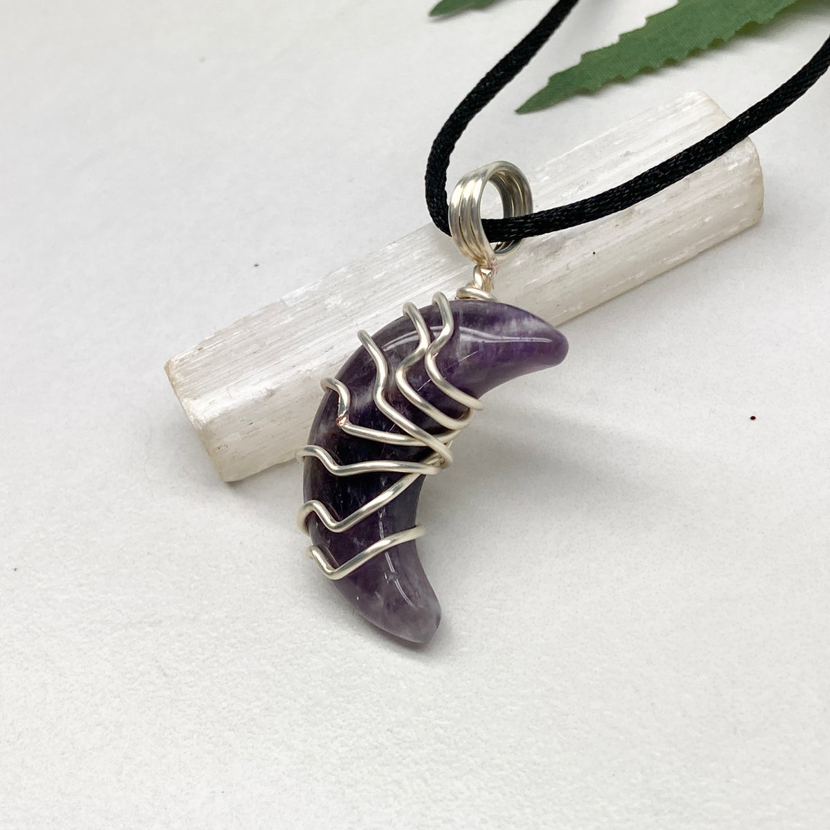 Purple Amethyst crescent moon necklace wrapped in silver-colored copper wire and hanging on a black chain. Handmade during the Moon in Leo.