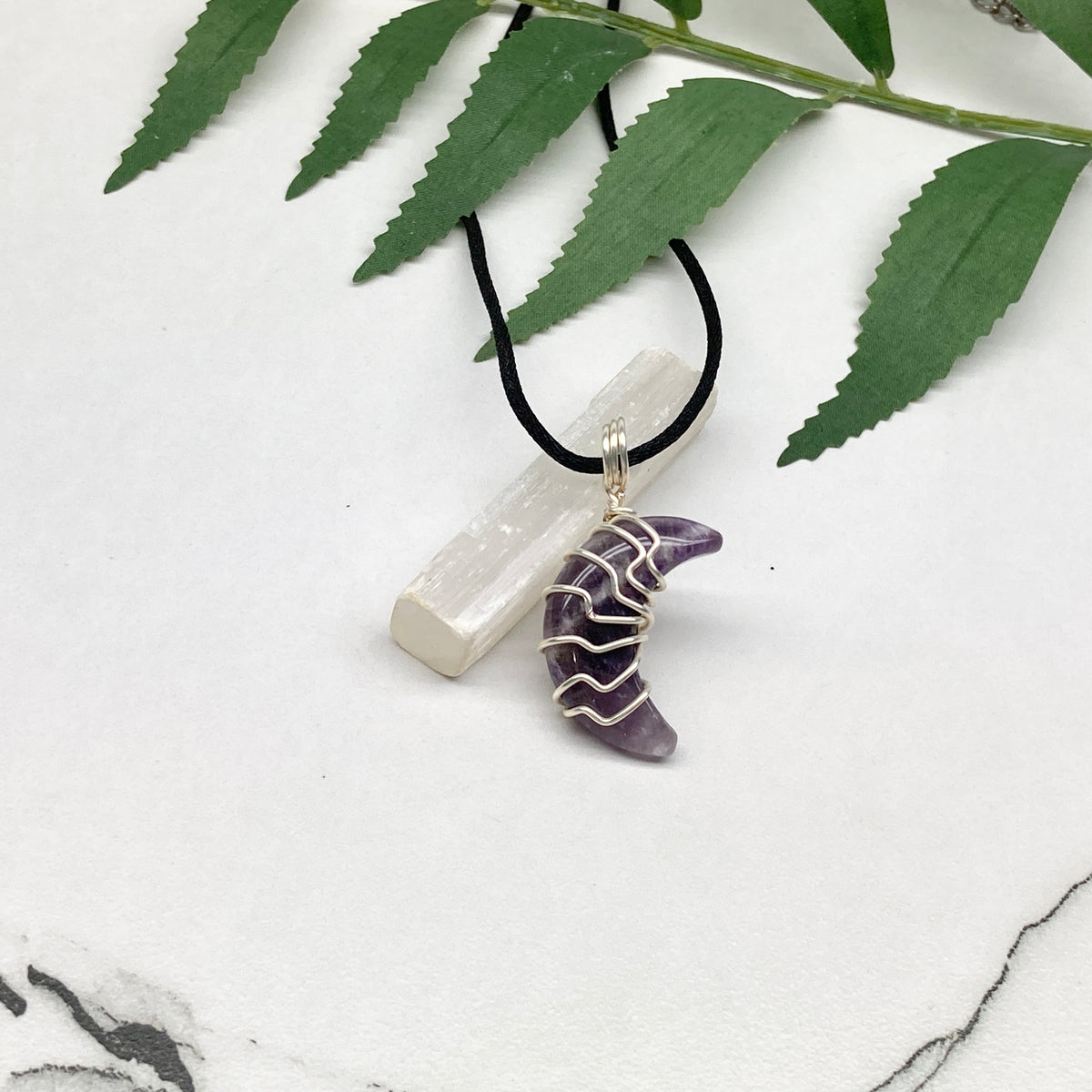 Purple Amethyst crescent moon necklace wrapped in silver-colored copper wire and hanging on a black chain. Handmade during the Moon in Leo.