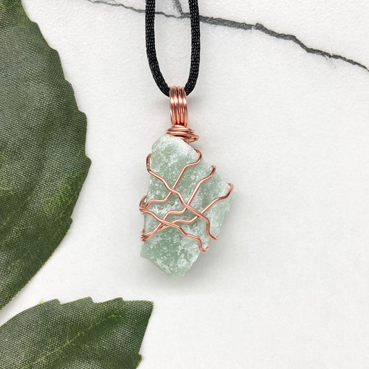 Green crystal pendant with copper wire wrapping on a black cord, placed on a marble surface with green leaves.